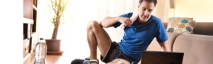 Man in workout clothes sitting on the floor at home, looking at a laptop, and touching his knee as if in pain.