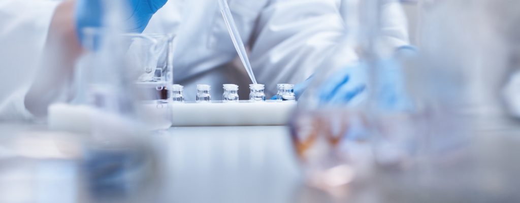 Scientist filling vials with solution through pipette in test tube rack. Chemist is examining medicine during scientific experiment. She is wearing gloves at pharmaceutical factory.