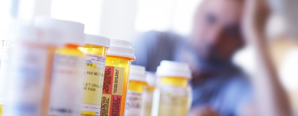 Medication Overload A large group of prescription medication bottles sit on a table in front of a distraught man who is leaning on his hand as he sits at his dining room table. The image is photographed with a very shallow depth of field with the focus being on the pill bottles in the foreground.